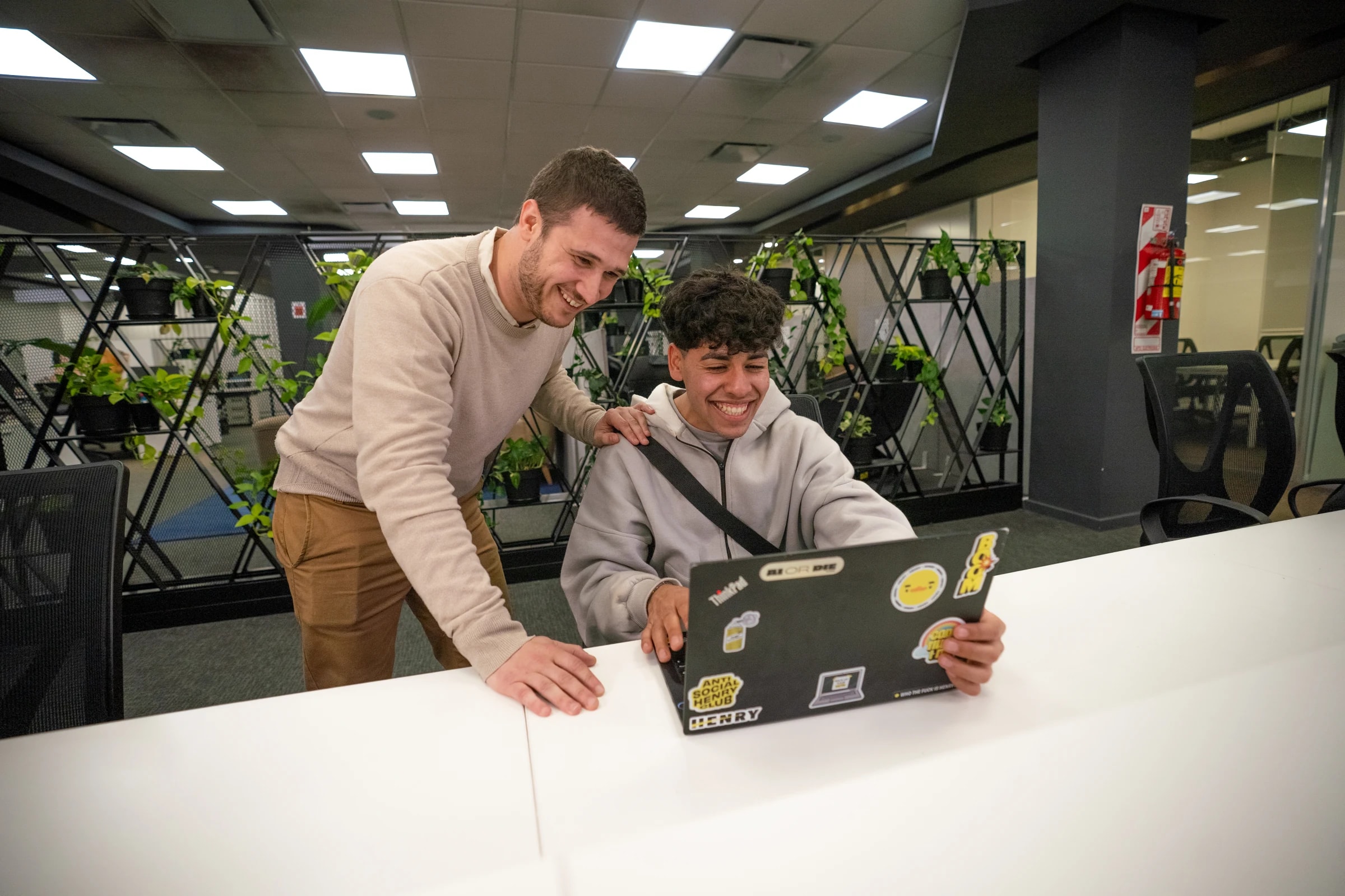 Dos personas sonríen frente a una notebook con stickers de Henry en un espacio de trabajo con plantas; una se apoya en el hombro de la otra