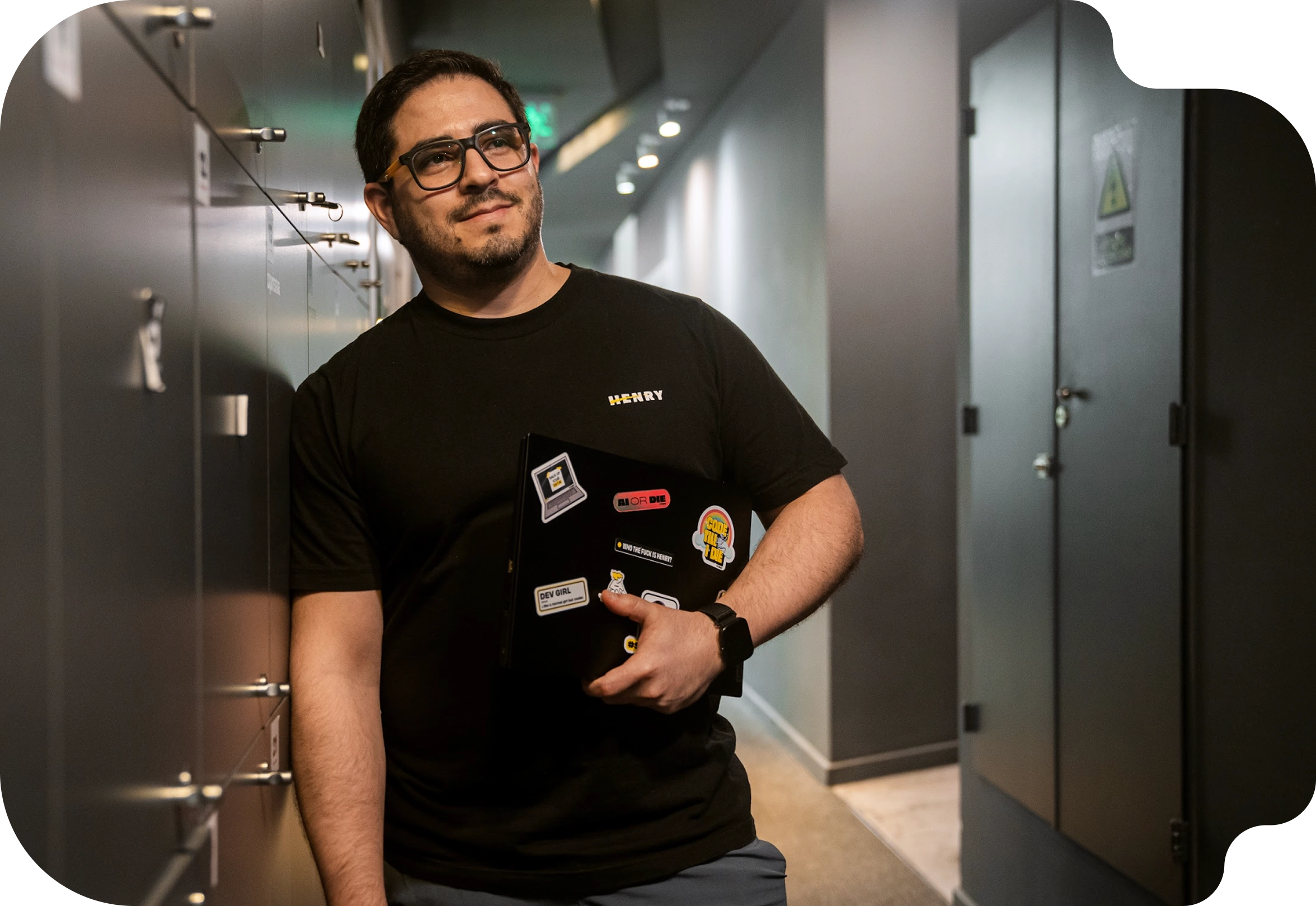 Un hombre de anteojos con la remera de Henry sostiene una notebook con stickers en un pasillo